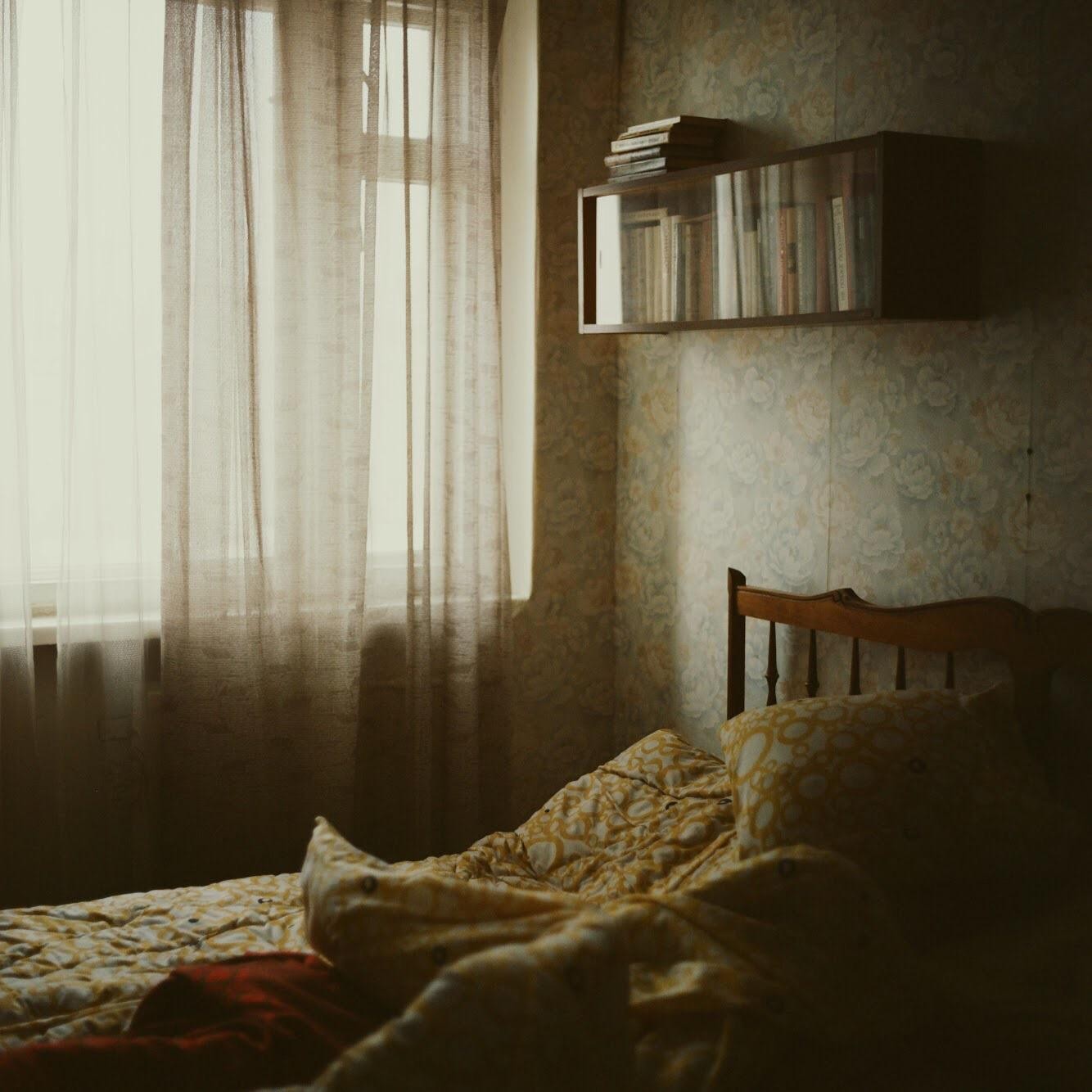 A bed with a patterned duvet and pillow sits next to a window with sheer curtains, with a bookshelf mounted on the wall above the headboard.