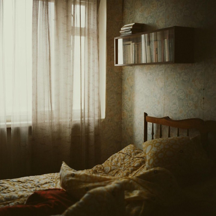 A bed with a patterned duvet and pillow sits next to a window with sheer curtains, with a bookshelf mounted on the wall above the headboard.
