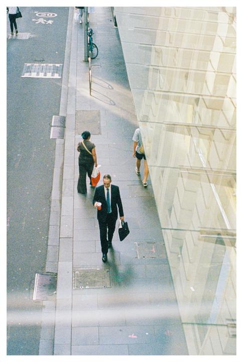 A man in a suit holding a cup and a bag walks on a city sidewalk next to a glass building.