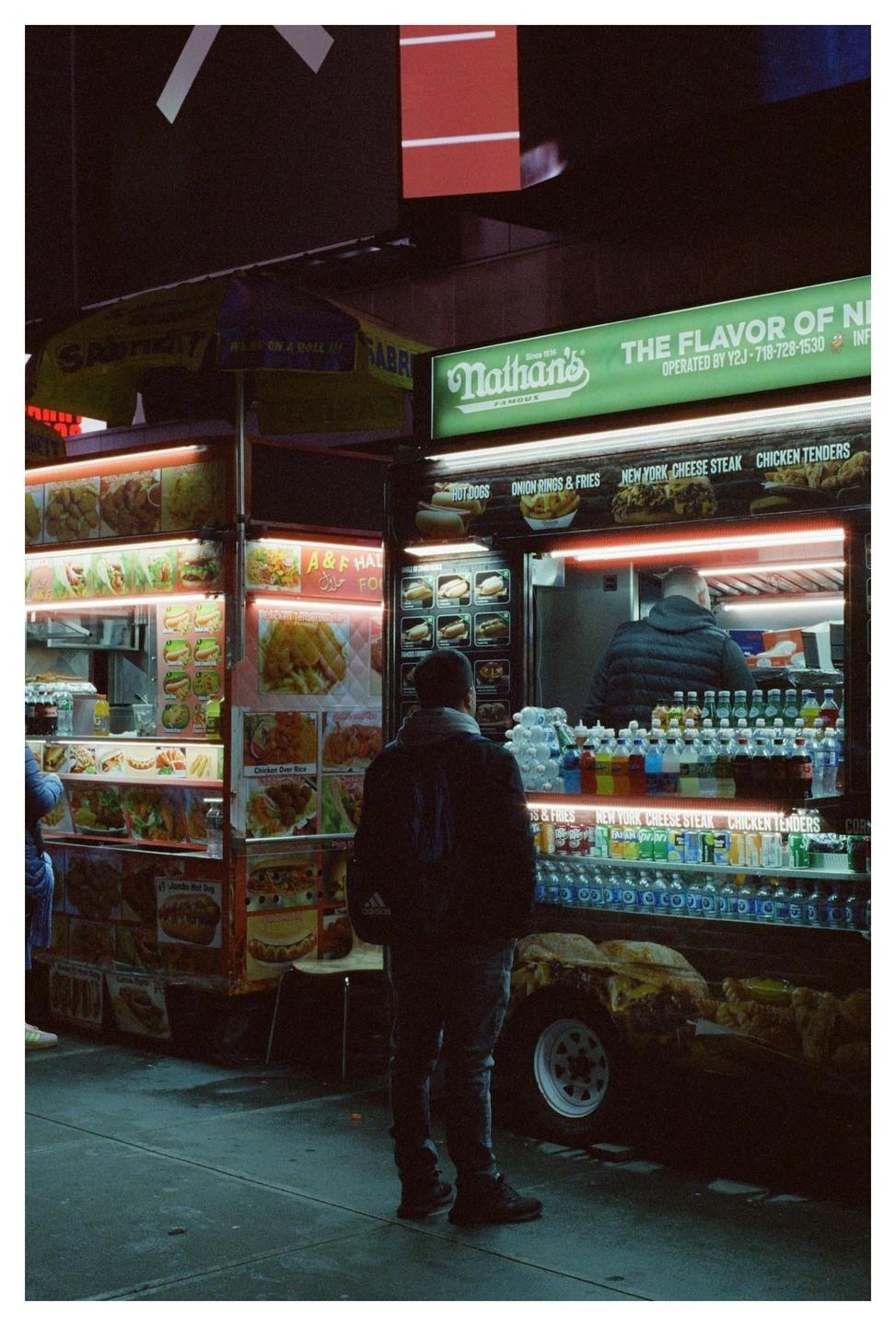 A man stands in front of a brightly lit food cart at night.