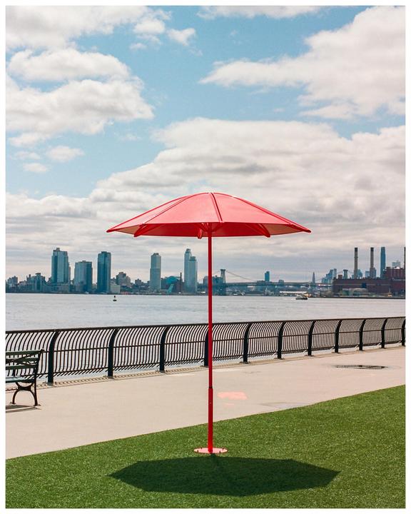 A red umbrella casts a shadow on a patch of grass next to a waterfront promenade with a city skyline in the background.