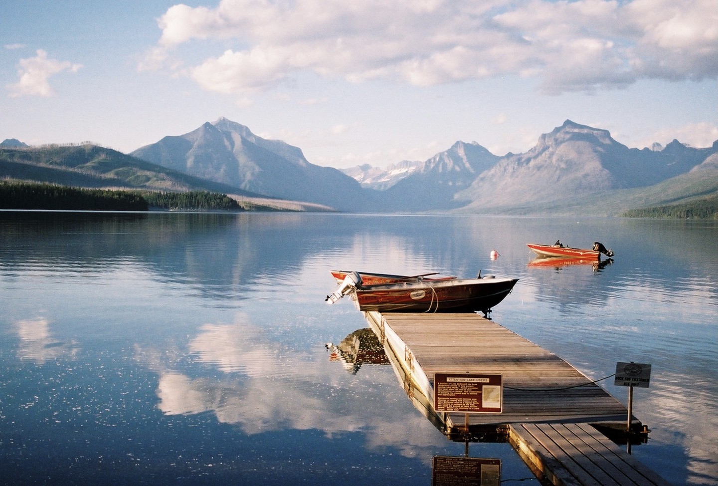 Two boats are docked at a wooden pier on a lake with mountains in the background.