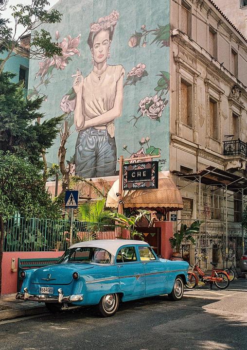 A blue vintage car is parked on a street next to a building with a large mural of a woman smoking.