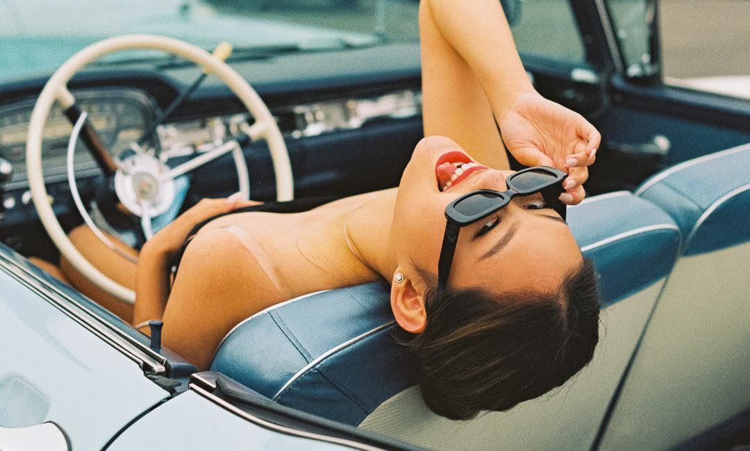 A woman with sunglasses and red lipstick is lying in a vintage car.