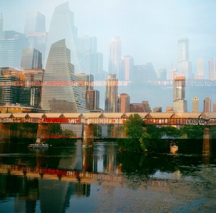 A double exposure shows a city skyline superimposed over a bridge with graffiti spanning a body of water.