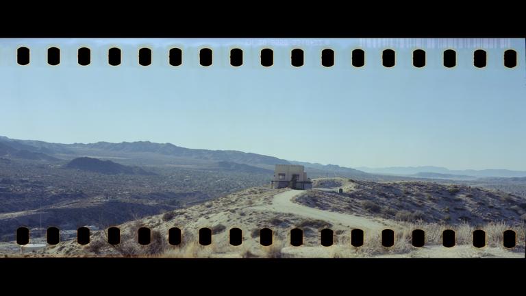 A building sits on a hill with a winding road leading to it, surrounded by a vast desert landscape under a clear sky.