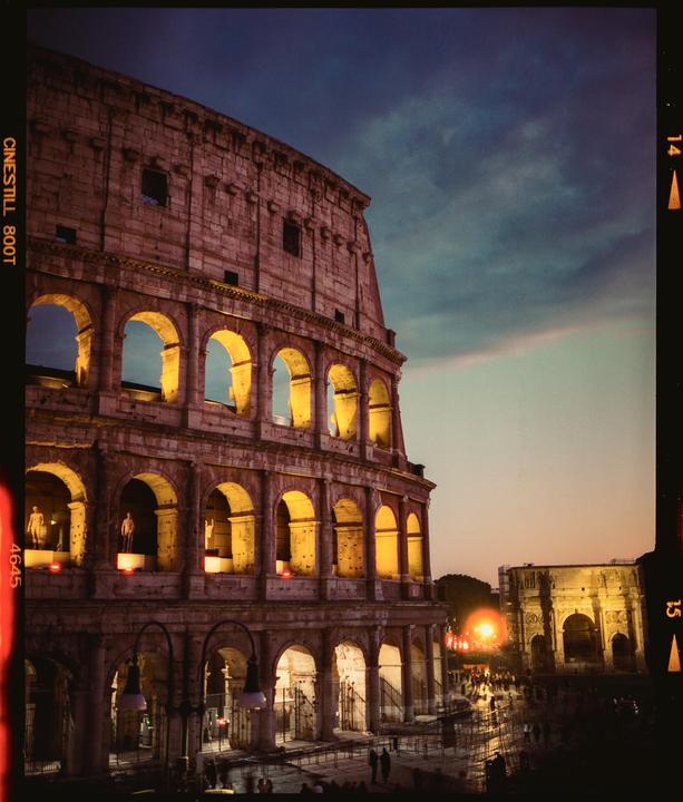 The Colosseum is illuminated at dusk with a cloudy sky overhead and people gathered in front.