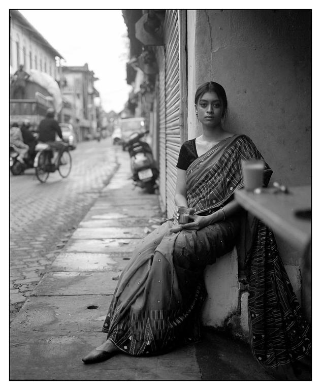 A woman in a sari sits on a ledge next to a street with buildings and vehicles.