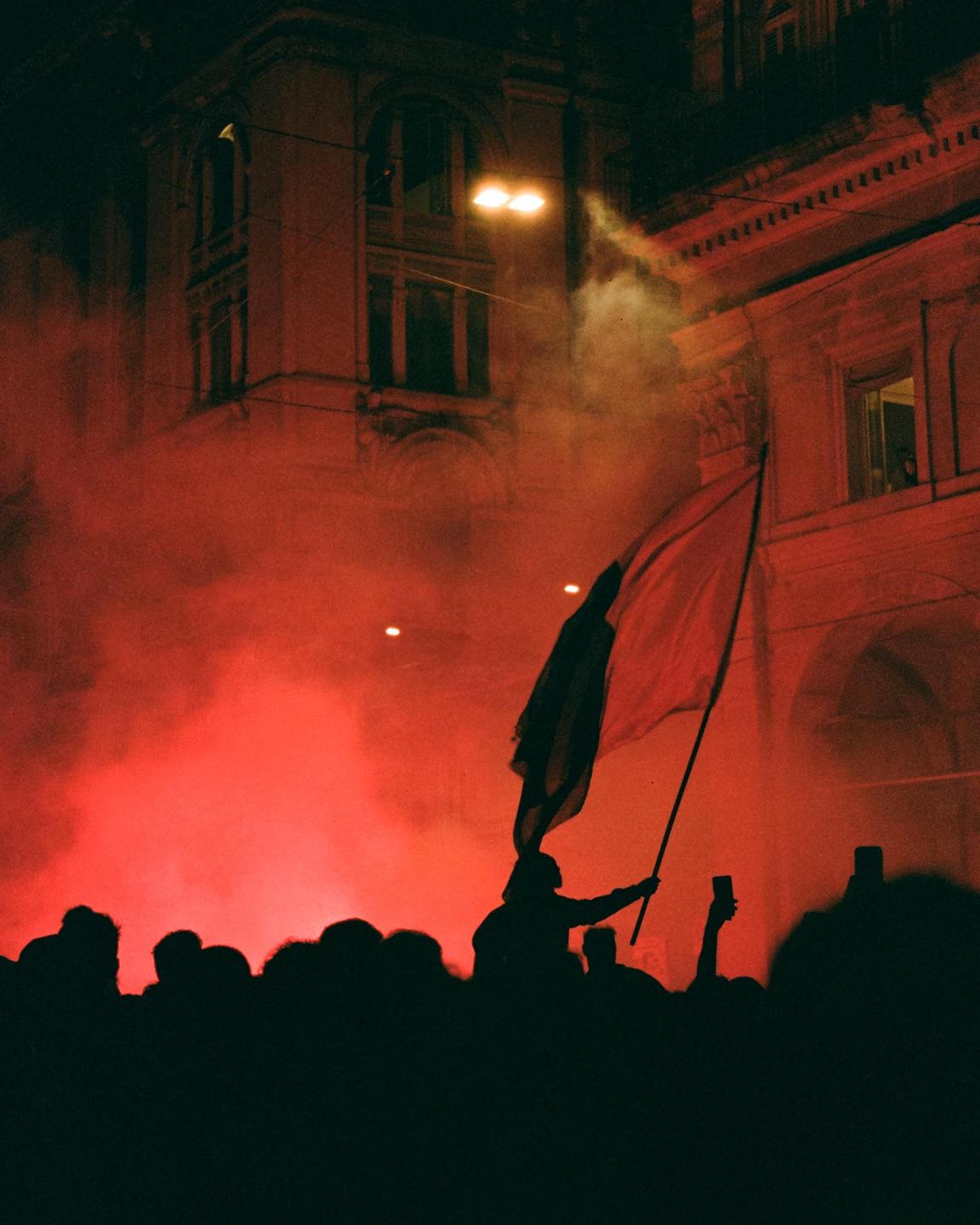 A crowd of people are silhouetted against a red smoky background with a building in the distance.