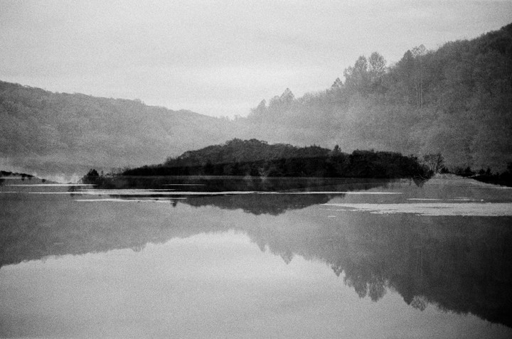 A body of water reflects a tree-covered island and hills in the background.