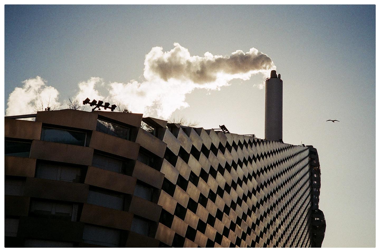 A tall smokestack emits a large cloud of smoke next to a building with a patterned facade.