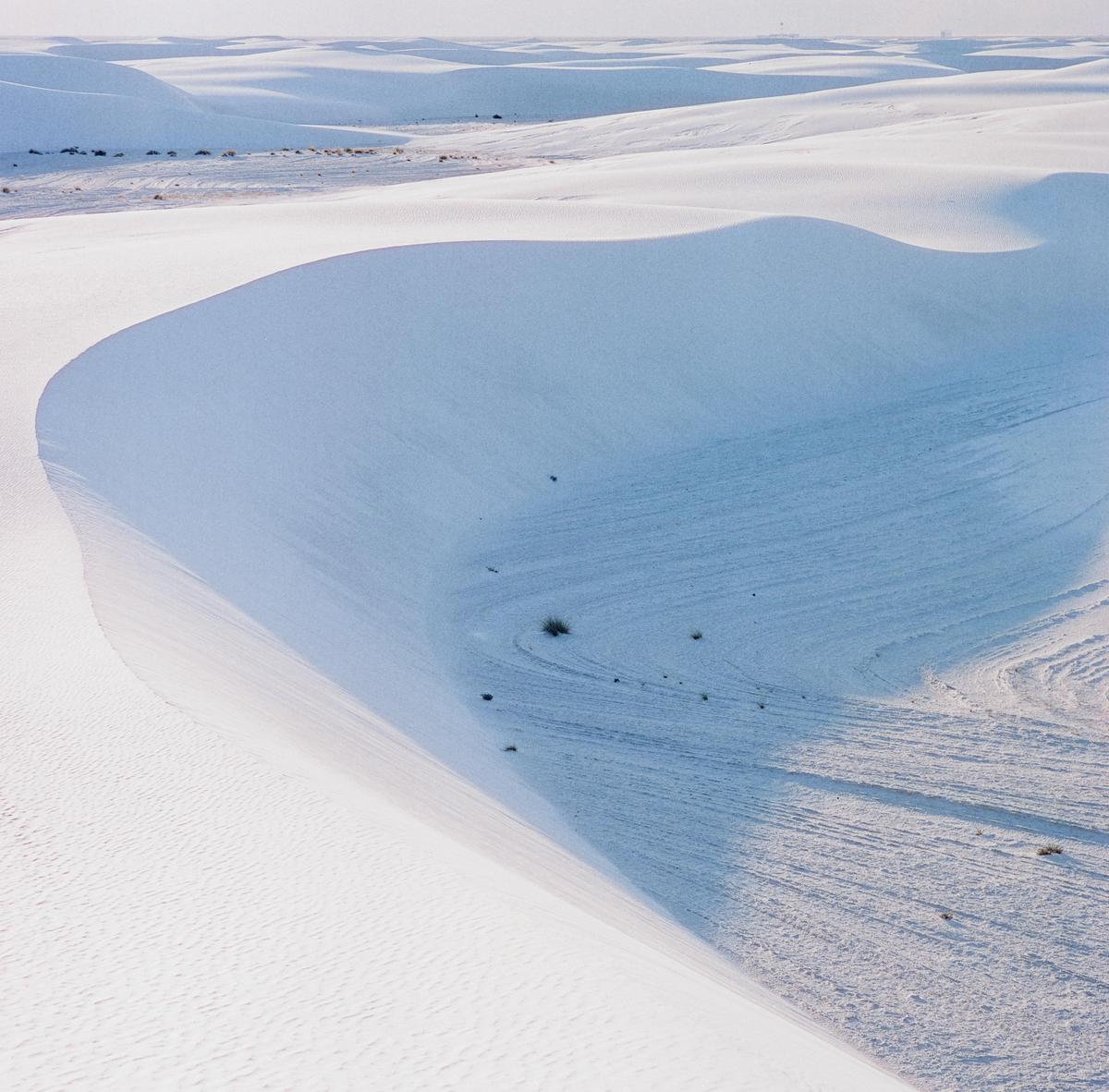 A vast desert landscape features white sand dunes under a clear sky.