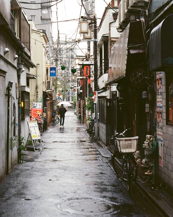 A man with an umbrella walks down a narrow, wet street lined with buildings and bicycles.