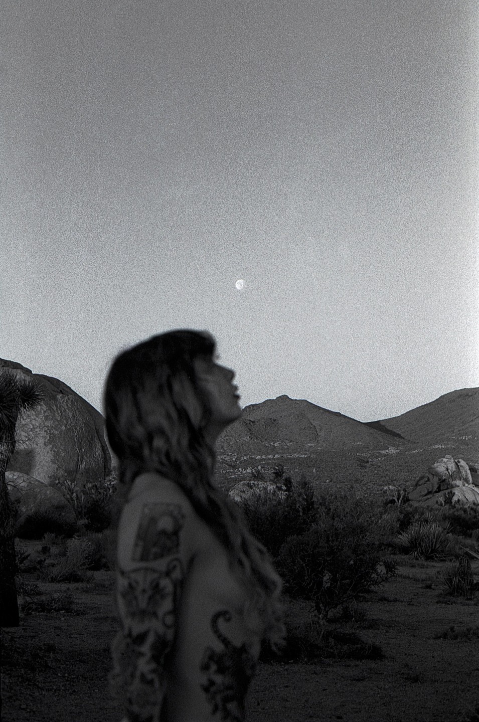 A woman with tattoos looks up at the moon in a desert landscape with mountains and rocks.