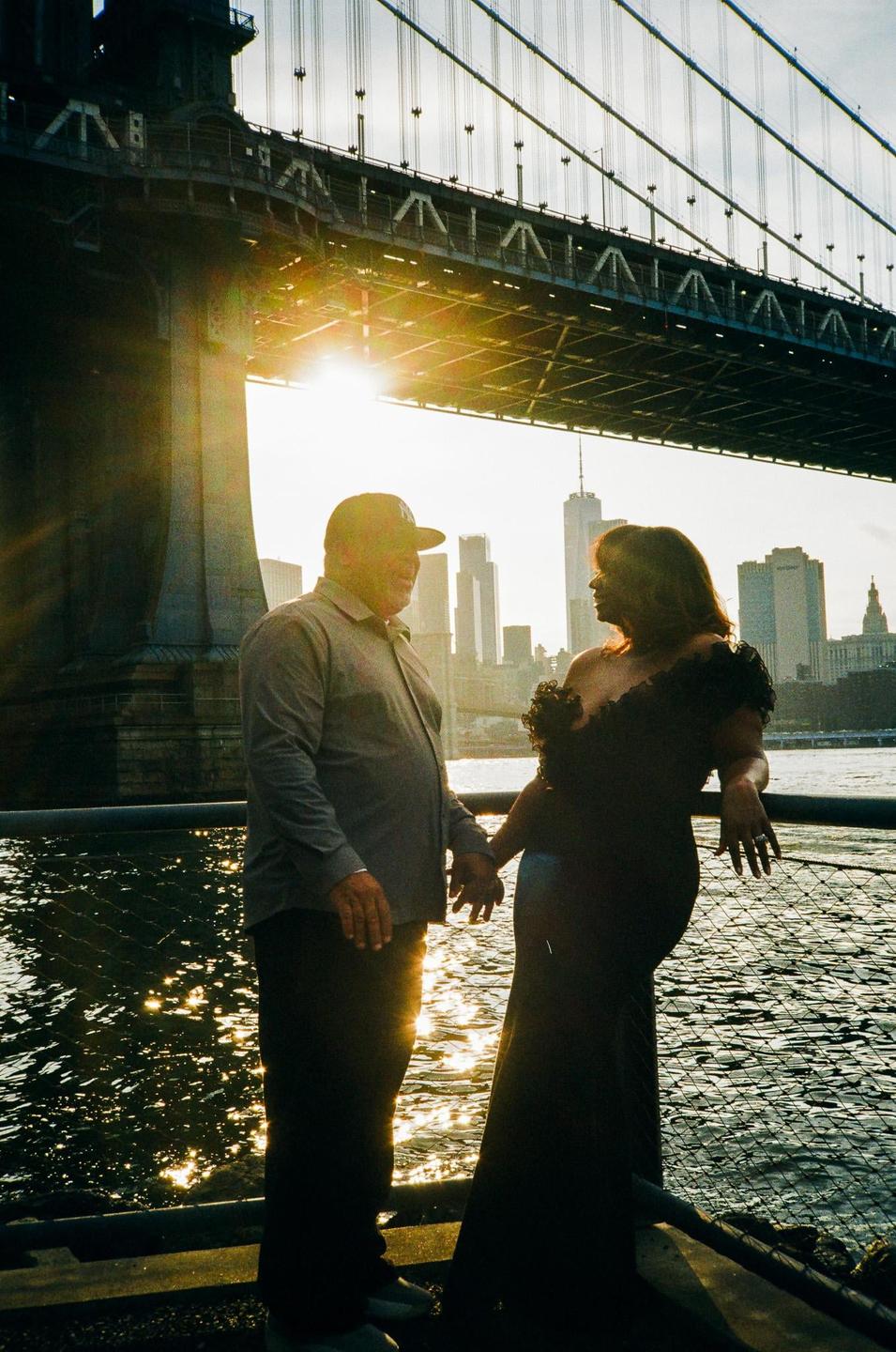 A couple stands holding hands by the water with a bridge and city skyline in the background at sunset.