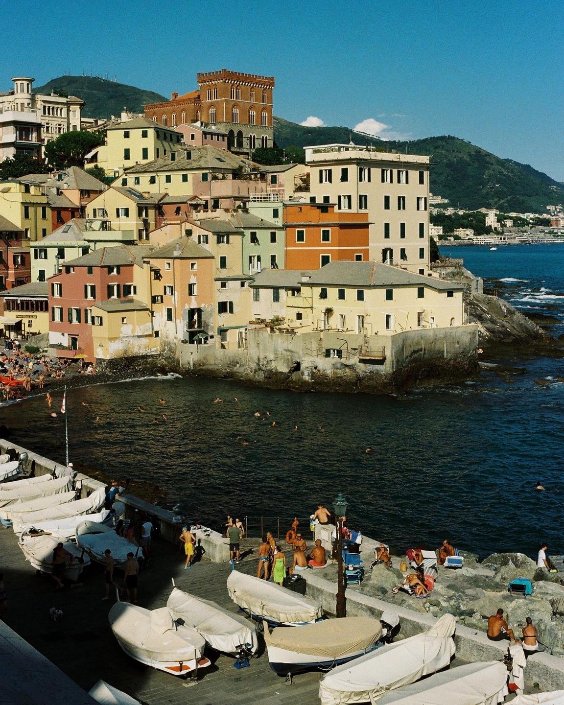 A coastal town with colorful buildings is nestled into a hillside overlooking a bay with people swimming and sunbathing.