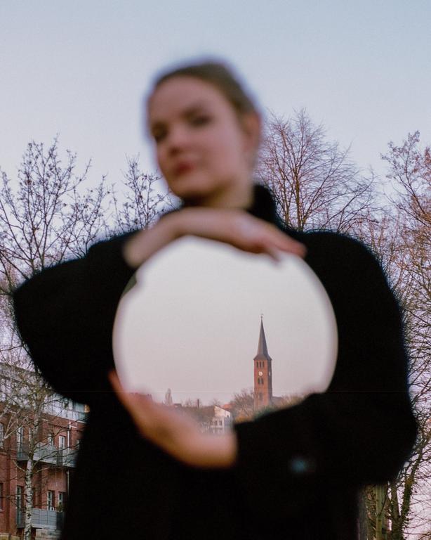 A woman holds a mirror reflecting a church steeple in a city.