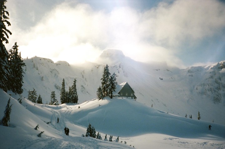 A small cabin sits on a snowy hill surrounded by pine trees and mountains under a cloudy sky.