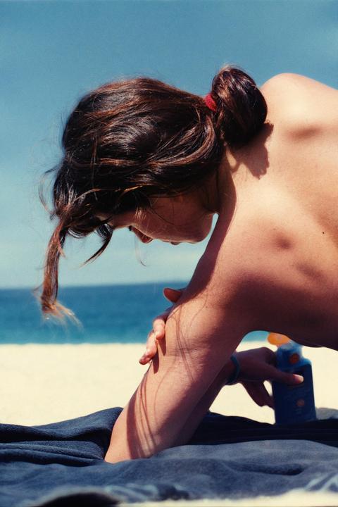 A woman applies sunscreen to her arm while lying on a towel on a beach.