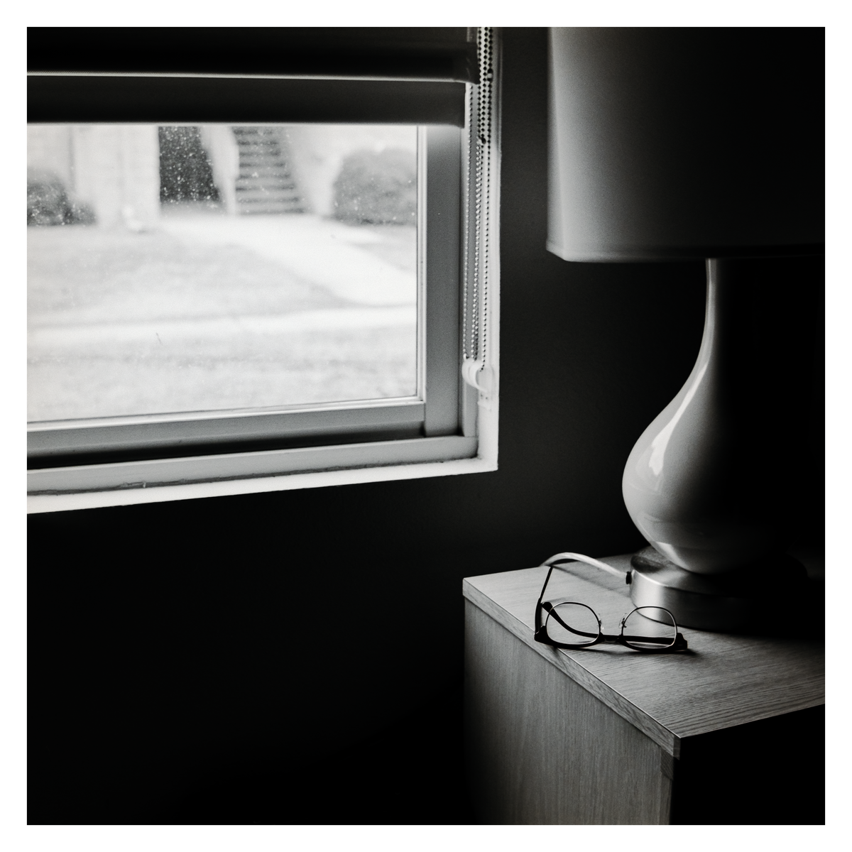 A pair of eyeglasses rests on a nightstand next to a lamp, with a window showing an outdoor view in the background.