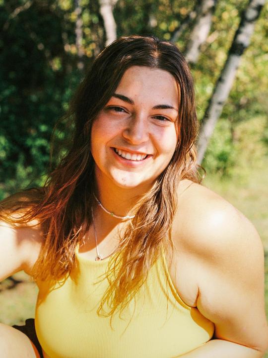 A woman with long hair and a yellow top smiles at the camera in a sunlit outdoor setting.