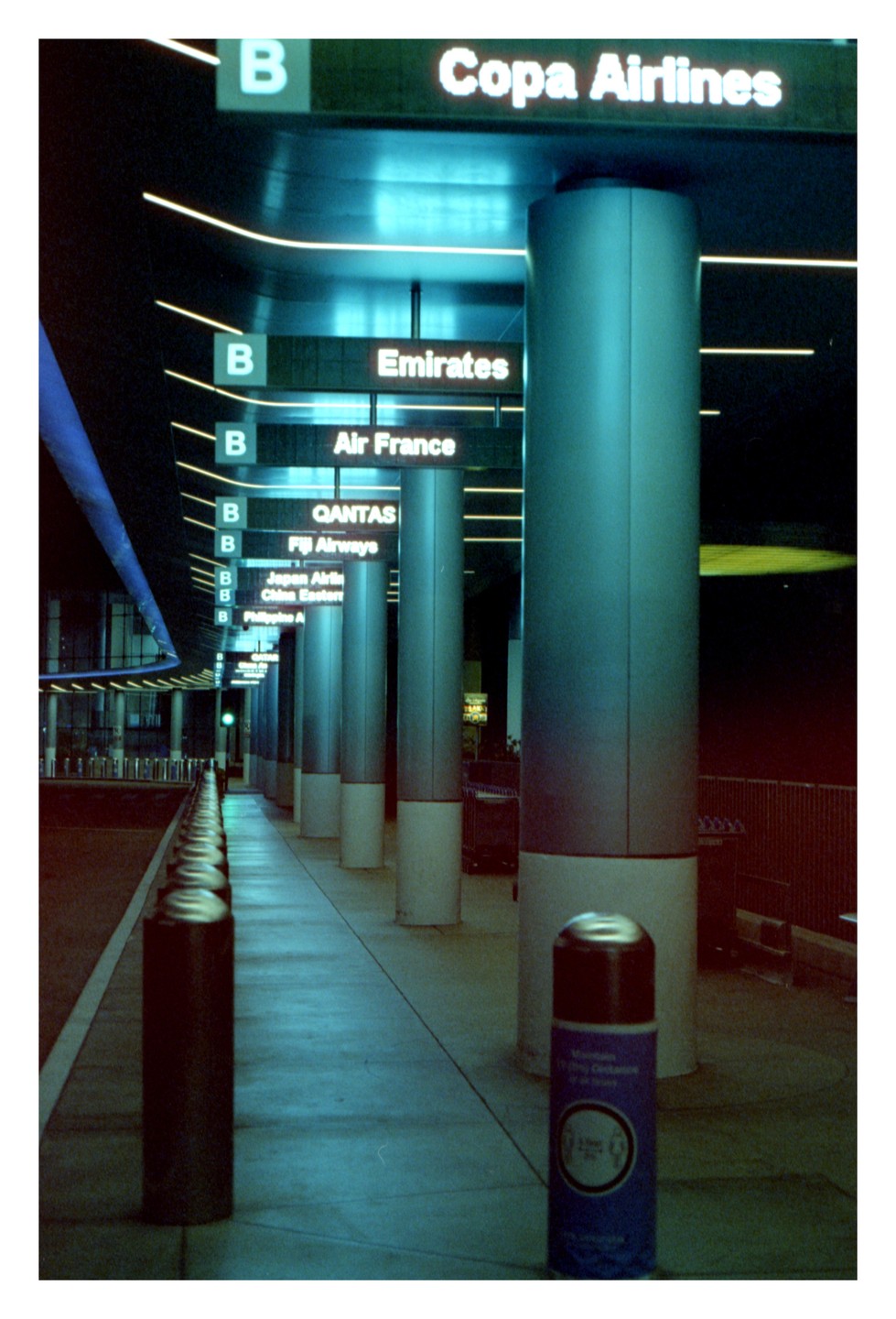 An airport terminal at night shows signs for various airlines and a row of bollards.