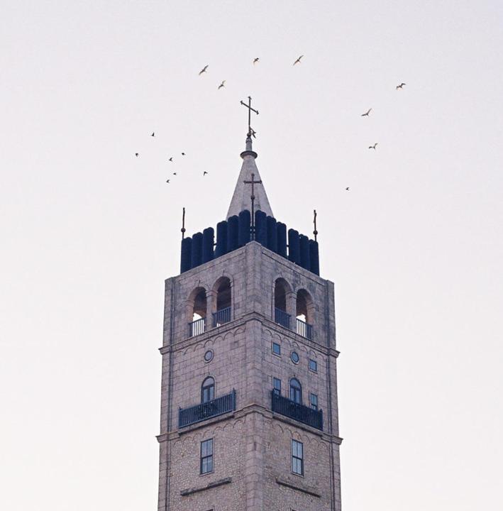 A stone church tower with a cross on top stands against a bright sky with birds flying around it.