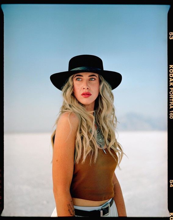 A woman with long blonde hair and a black hat looks to the side on a salt flat.