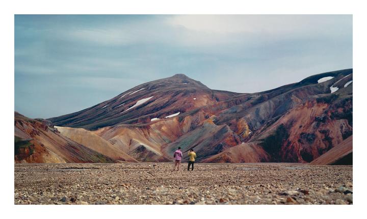 Two people stand on a rocky plain looking towards colorful mountains with patches of snow.