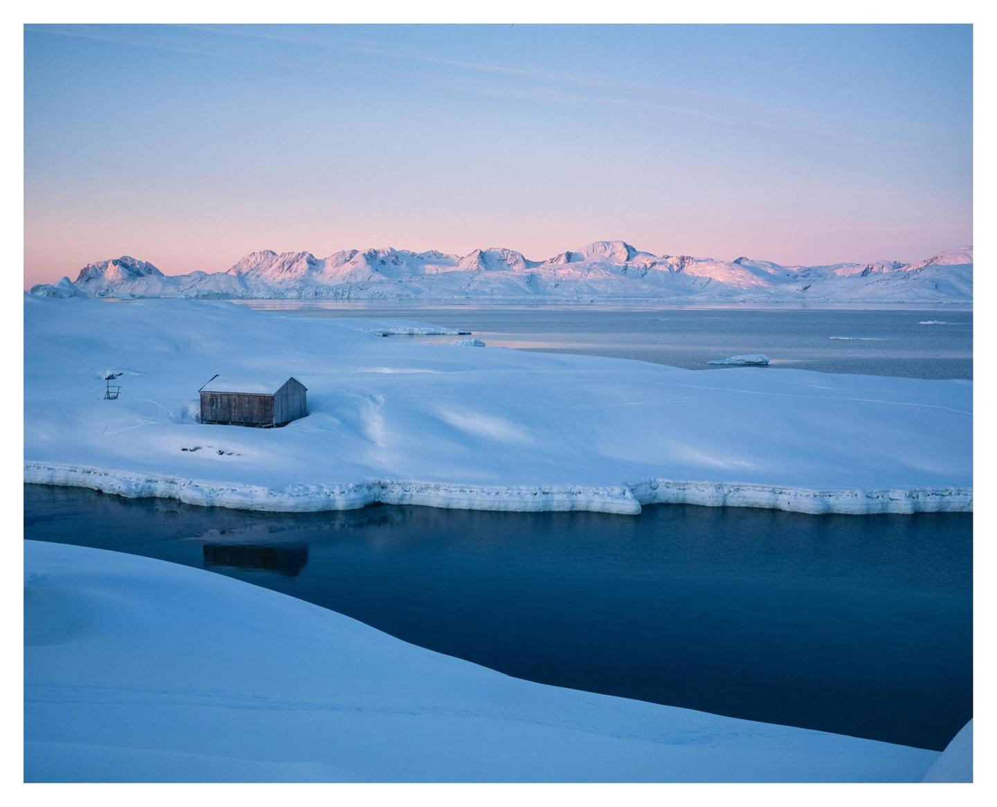 A small wooden cabin sits on a snow-covered bank next to a body of water, with mountains in the distance under a pink and blue sky.
