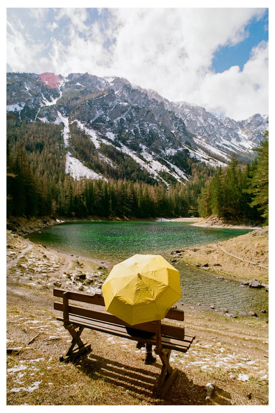 A person sits on a bench with a yellow umbrella, facing a green lake surrounded by a forest and snow-capped mountains under a cloudy sky.