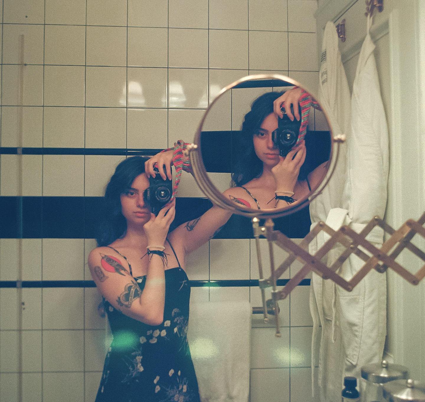 A woman in a floral dress takes a mirror selfie in a tiled bathroom, holding a camera to her face.