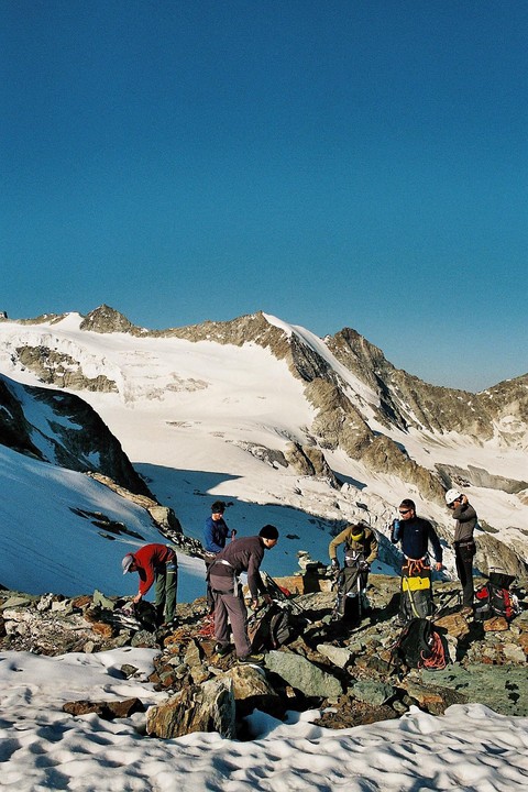 A group of men are preparing their gear on a rocky, snowy mountain with a large snow-covered peak in the background.