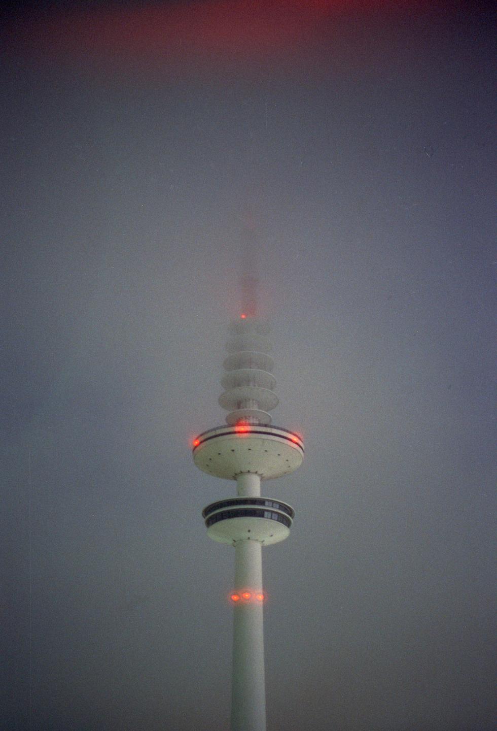 A tall, white television tower with red lights is partially obscured by fog.