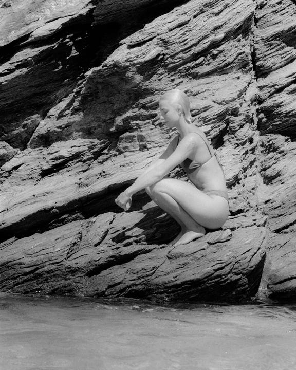 A woman in a swimsuit crouches on a rock next to water with a large rock formation behind her.