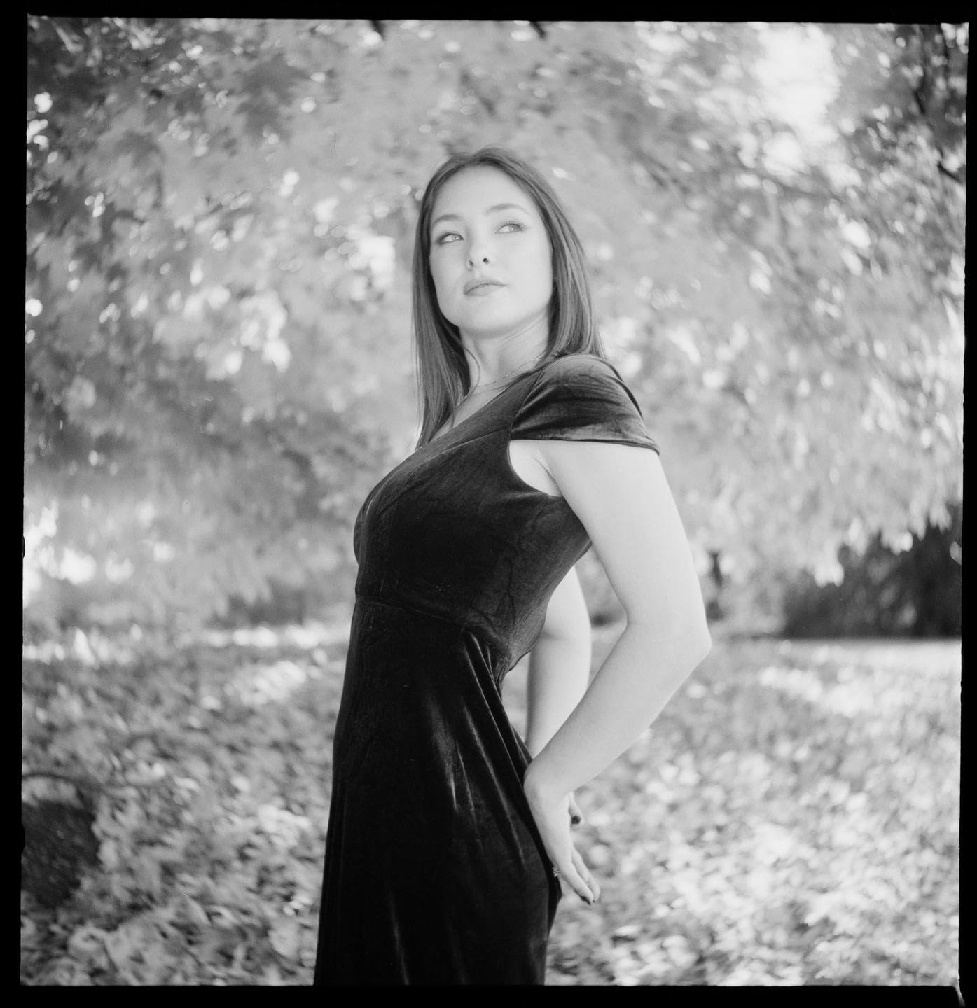 A woman in a dress stands in a park with trees and fallen leaves in the background.