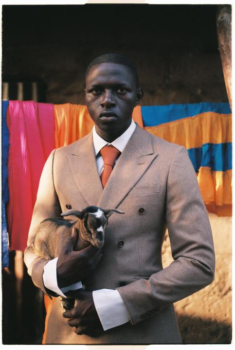A man in a suit holds a baby goat in front of colorful fabric.