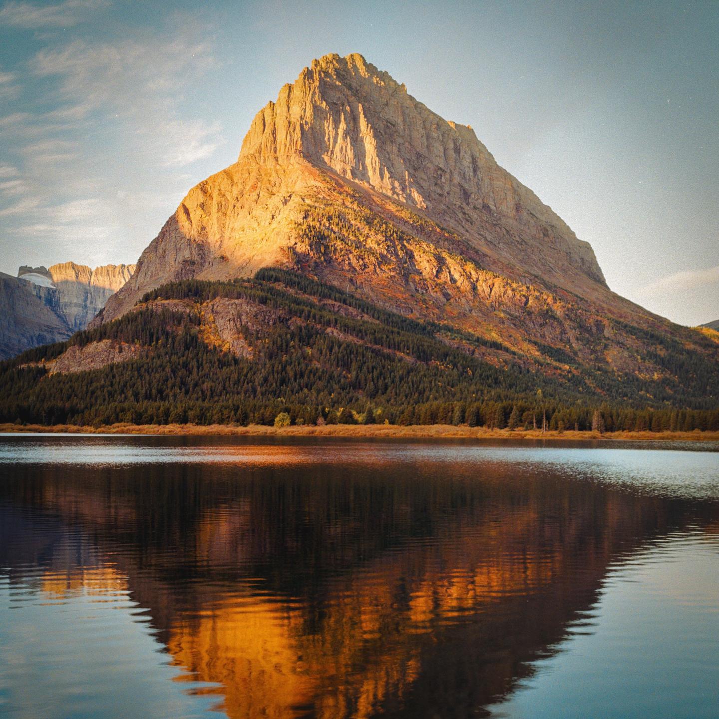 A large mountain with trees on its lower slopes is reflected in a lake under a bright sky.