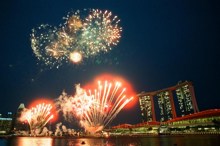 Fireworks explode over a body of water with city buildings in the background at night.