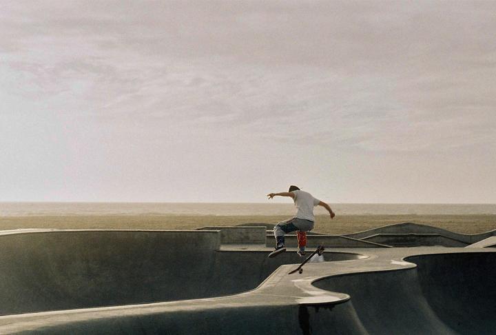 A man on a skateboard is captured mid-air over a skatepark with the ocean in the background.