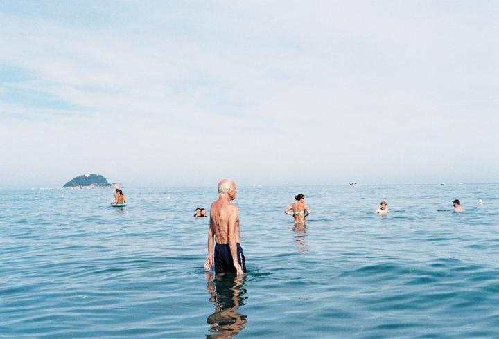 A man stands in the ocean with other people swimming in the background and an island in the distance.