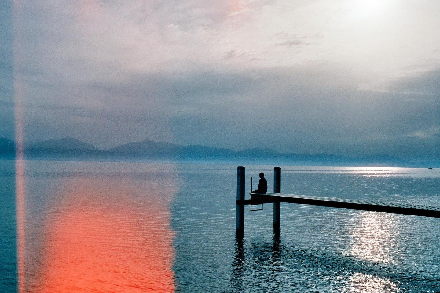 A person sits on a dock over a lake with mountains in the distance under a cloudy sky.