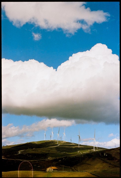 A large cloud hangs over a hill with wind turbines and a small house.