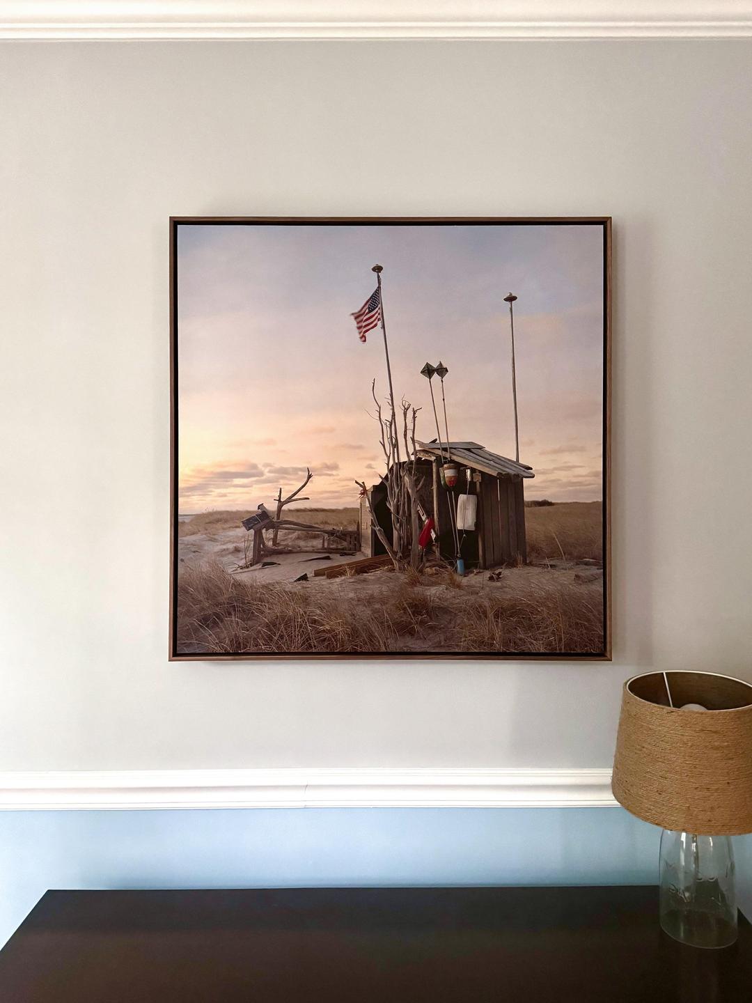 A framed print of a wooden shack with an American flag on a sandy beach is displayed on a wall above a dark table with a lamp.