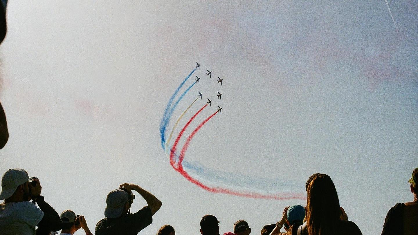 A group of people watch an airshow with several planes flying in formation and leaving red, white, and blue smoke trails in the sky.