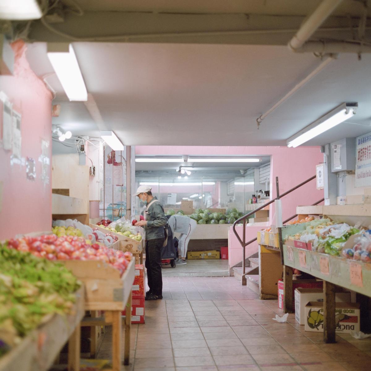 A man in a cap and mask selects produce from a wooden bin in a pastel pink supermarket.
