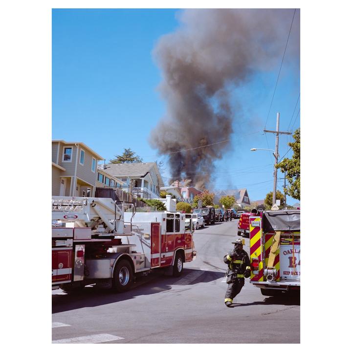 A firefighter runs down a street with fire trucks and a large plume of smoke rising from houses in the background.