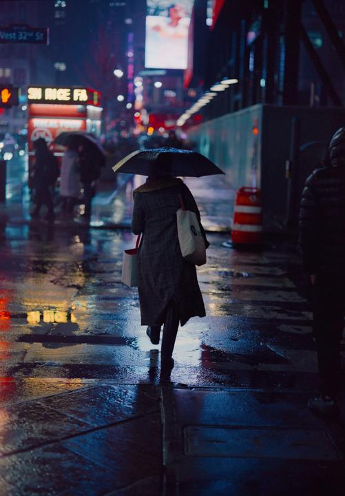 A woman with an umbrella walks down a wet street at night, with reflections of neon signs on the pavement.