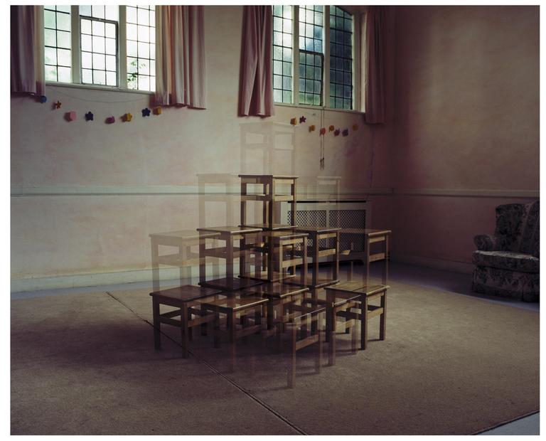 A multiple exposure shows a pyramid of wooden stools in a room with windows and pink curtains.