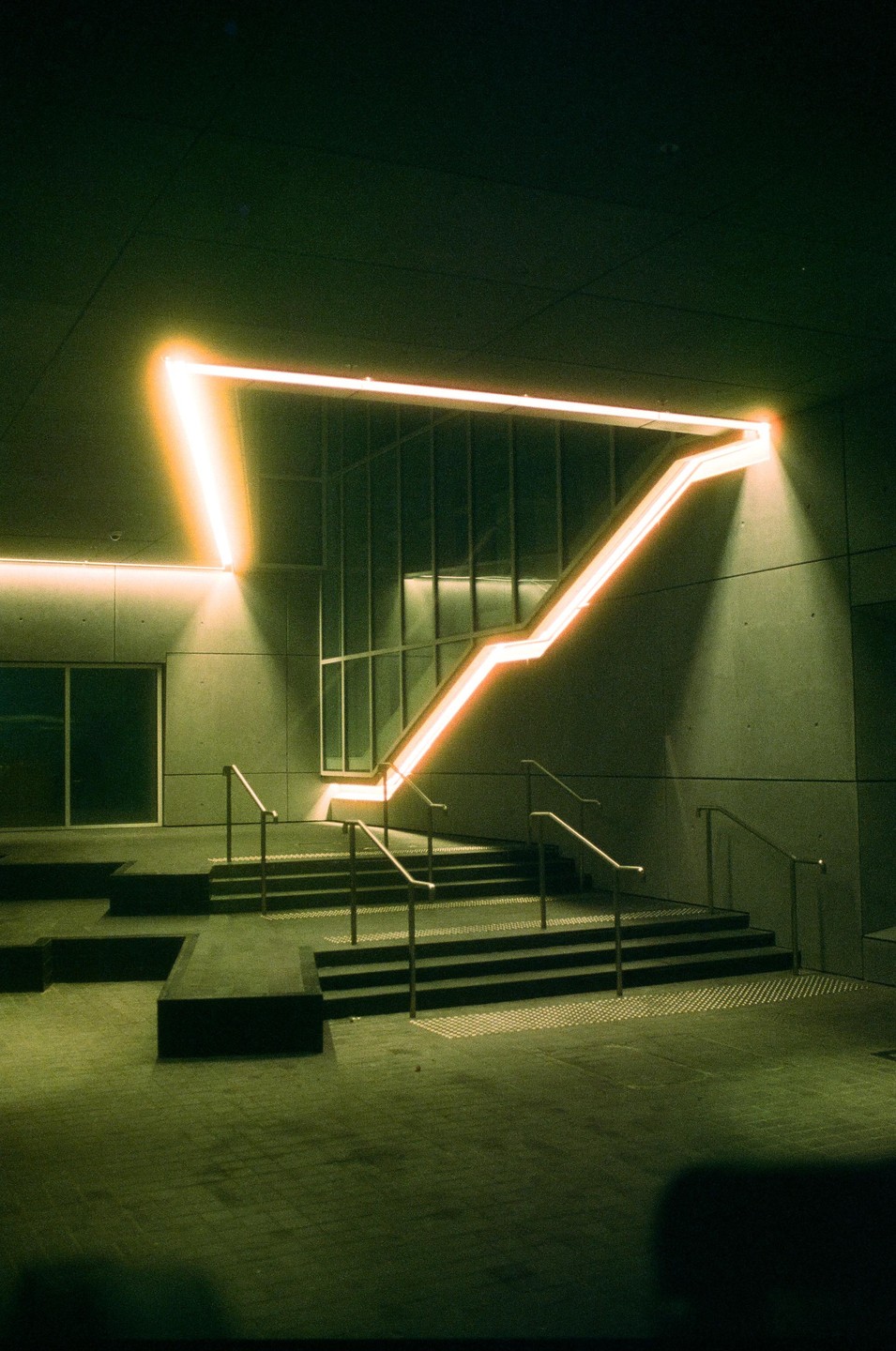 An illuminated orange neon light in the shape of a triangle is mounted on a concrete wall above a set of stairs with metal railings.